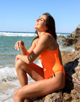 Side view of model on a rock wearing JAYD Swimwear athletic one piece Apsley in Neon Orange.