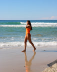 Side view of model walking on a beach wearing JAYD Swimwear athletic one piece Apsley in Neon Orange.