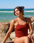 Front view of model sitting on a rock wearing JAYD Swimwear athletic one piece Isla in Cinnamon.