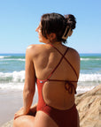 Back strap view of model sitting on a rock wearing JAYD Swimwear athletic one piece Isla in Cinnamon.