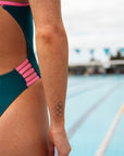 Close hip cut out view of model standing near a pool wearing JAYD Swimwear athletic Lennox one piece in cove.