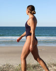 Side view of model walking on sand wearing JAYD Swimwear cheeky one piece Zimmy in Acai.