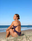 Side view of model sitting on sand wearing JAYD Swimwear cheeky one piece Zimmy in Acai.