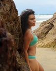 Side view of model leaning against a rock wearing JAYD Swimwear athletic Torquay training bikini top in Matcha green.