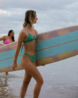 Model walking with surfboard in JAYD Swimwear Cali athletic bikini bottoms in Matcha Green by JAYD Swimwear