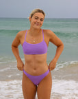 Front view of a female athlete in a JAYD Swimwear Torquay bikini top in lilac haze, standing on a beach with waves in the background.