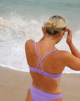 Close back strap view of a female athlete in a JAYD Swimwear Torquay bikini top in lilac haze, standing on a beach with waves in the background.