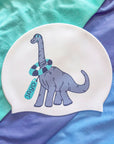 A white swim cap featuring a cartoon Brachycephalus dinosaur in gray with a teal and black striped swim ring and a teal tag that reads 'JAYD', laid flat on a draped background of teal and blue fabric.