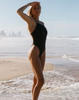Woman in a cacao brown athletic one piece standing on a beach with ocean waves and skyline in the background
