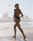 Athlete in a cacao brown fixed back one piece walking on a beach with trees in the background.