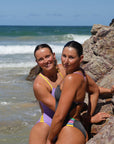 Two models wearing JAYD Lennox One Piece swimsuits on a sunny day on the beach with the ocean waves breaking in the background.