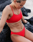 Side view of surf lifesaving athlete Chelsea Woods in a JAYD Swimwear Torquay bikini top in Ruby, sitting on a rock down by the beach.