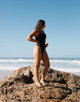 Woman in a black JAYD one piece standing on a rock at the beach with clear blue sky. She is wearing the Albi style