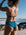 Woman in a Nordic blue full-coverage Connie bikini bottom, standing on a beach with ocean view.