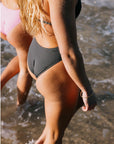 Woman wearing a Tallow One Piece Swimsuit in our beautiful dark grey shade, Carbon. Standing in shallow water at the beach.