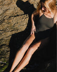JAYD Swimwear Tallow one piece in Carbon grey colour, Brodee is sitting on the rocks.