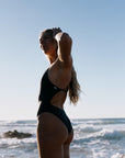 Woman in the TALLOW One Piece swimsuit in our Carbon grey shade, standing on a beach with ocean waves in the background
