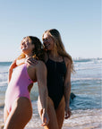 Sisterly love in the TALLOW One Piece Swimsuit - featuring athletes, Brodee and Morgan, standing on a beach with ocean waves in the background