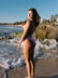 The TALLOW swim one piece, in Fairy Floss pink, worn by Morgan standing on a beach with ocean waves and rocks in the background