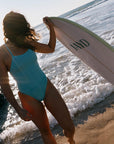 Ocean athlete, Madi, wearing the Tallow One Piece in Mykonos, holding a surfboard on a beach with waves.