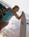 Woman in a light blue (shade: Mykonos) Tallow One Piece swimsuit holding a surfboard with 'JAYD' branding on a beach.