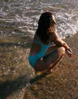 The TALLOW fuller coverage one piece swimsuit in Mykonos Blue, worn by Madi sitting on a beach with waves.