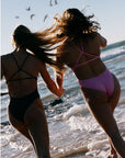 Two women in TALLOW fuller coverage fixed-back swimsuits, running on a beach with waves and birds in the background.
