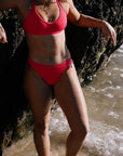 Madi wears the Connie full-coverage bottom in Fiesta, standing by a rocky shoreline.