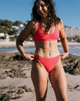 Woman in a "Fiesta" red bikini, featuring the JAYD Connie Bottom and Tazzie top, standing on a beach with a scenic background