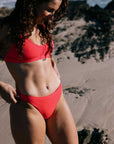 Athlete Madi Armstrong, in a "Fiesta" red bikini standing on a beach with sand and rocks in the background