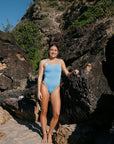 Lucy wears the athletic one piece called Albi, she is in the Breeze colour hue and is standing, leaning against the rocks