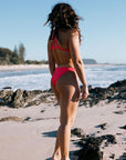 Madi wears a Fiesta bikini bottom offering mid coverage, called Cali, standing on a beach with ocean and sky in the background
