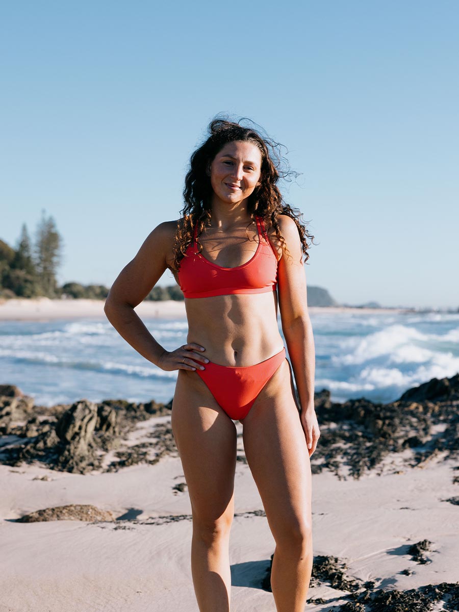 Madi wears her Fiesta colours bikini standing on a beach with ocean waves in the background. She is wearing our mid coverage Cali bottoms.