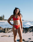 Madi wears her Fiesta colours bikini standing on a beach with ocean waves in the background. She is wearing our mid coverage Cali bottoms.