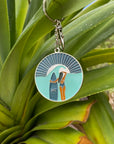 JAYD Swimwear keychain featuring a woman with a surfboard staring into the distant sunset on a beach, hanging from a plant.