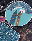 JAYD Swimwear keychain featuring a woman with a surfboard staring into the distant sunset on a beach, sitting on a rock with other colourful keychain designs.