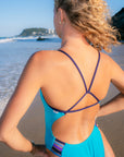 JAYD Swimwear model on a beach wearing the Lennox one-piece swimsuit in atlantic with dark purple acai crossover back straps, shown from behind.