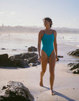 Model Sophie walking along a beach wearing the JAYD Swimwear Nordic Lennox one piece with contrasting navy straps.