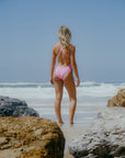 JAYD model standing on a beach in the fairy floss Levanto one piece, facing the ocean with the suit's wide cross-back straps on display.