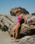 JAYD model Adri leaning against a rock at the beach wearing the Levanto one piece swimsuit in Orchid.