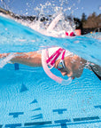 Underwater close-up of a swimmer wearing a pink FINIS Stability Snorkel snorkel and goggles in a blue swimming pool, with water splashing above the surface.