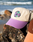 Salty Summer embroidery design on the cream JAYD Swimwear baseball cap with purple brim, sitting on a rock with the ocean in the distance.