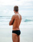 JAYD Swimwear model wearing the black Ramshark men's swim bottoms, showcasing the comfortable moderate coverage fit, staring off into the distance at the beach.