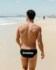 Man walking on a beach wearing black swim briefs with customised 'MOONERS' branding.