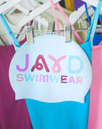 White swim cap with squiggly colourful 'JAYD Swimwear' branding on a rack with complimentary sangria, fairy floss and cancun one pieces.