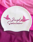 A white swim cap with pink printed graphics of two hummingbirds and the text 'Jayd Swimwear', displayed on a pink and magenta fabric background.