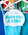White swimcap with 'Swim for a cure' copy and Cure Cancer x JAYD logo hanging with blue JAYD swimsuits.