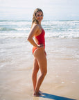 Athlete Hunter wearing the ruby Tallow one-piece swimsuit stands on a sandy beach with ocean waves in the background.
