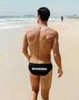 Man walking on a beach wearing black swim briefs with customised 'MOONERS' branding.