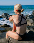JAYD Swimwear model with blonde hair in a bun sits on rocks by the ocean, viewed from behind, wearing the tick strap cross-back cacao Vienna bikini top. The background features a rocky shoreline and a blue ocean under a clear sky.